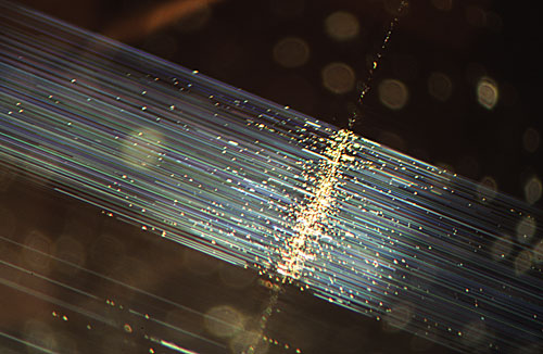 Figure 2. When fiber-optic illumination is used on the same chrysoberyl, bundles of fine light-scattering silky needles are seen extending at 90˚ from the linear growth band which is also more clearly detailed now. With fiber optics a tremendous amount of detail is gained. Magnified 10×. Photomicrograph © John I. Koivula, microWorld of Gems.